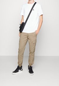 Vit t-shirt med kort ärm och svart logotyp, beige cargobyxor med sidofickor och svarta högskor med vita detaljer.