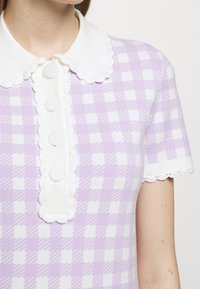 Kobieta ubrana w lawendową i białą koszulę w kratę z krótkim rękawem, z falbaniastym białym kołnierzykiem i guzikami z przodu.