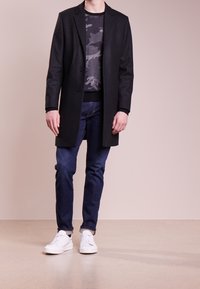 Cappotto lungo in lana nera con taglio dritto, abbinato a un maglione a fantasia camouflage, jeans scuri e sneaker bianche su uno sfondo neutro.
