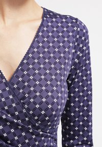 Námořnická modrá zavinovací blůza z hladké tkaniny, s diamantovým vzorem a malými bílými květinovými detaily, a s vykrojeným výstřihem.