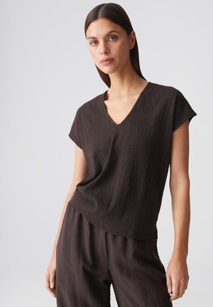 Donna con capelli scuri e lisci che indossa una maglia nera a maniche corte con scollo a V e pantaloni neri larghi su uno sfondo chiaro uniforme.