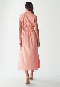 Moteris su ilgais plaukais, vilkinti rožinę be rankovių midi suknelę su elastingu juosmeniu, koja žengia atgal ant balto fono, avinti smėlio spalvos sandalus.