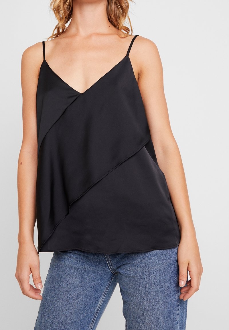 Svart satin kamisol med djup V-ringning och asymmetrisk lagerdesign. Tunna band och en mjuk textur; kombinerad med blå denimjeans.