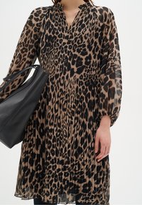 Femme portant une robe plissée à imprimé léopard avec des manches longues transparentes, tenant un grand sac fourre-tout en cuir noir sur un fond blanc.