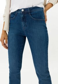 Jeans in denim blu scuro con un design aderente, stile a cinque tasche, chiusura a bottoni e dettagli di cucitura sottili su tasche e cuciture.
