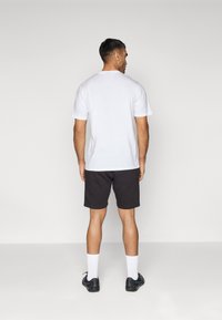 Camiseta blanca de algodón, de mangas cortas, combinada con pantalones cortos negros. El modelo lleva calcetines blancos y zapatos negros, visto desde atrás.