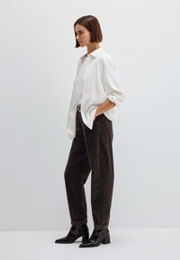 Camicia bianca a maniche lunghe con bottoni sopra una maglietta bianca, abbinata a pantaloni larghi a vita alta di colore marrone scuro e stivaletti neri con tacco.