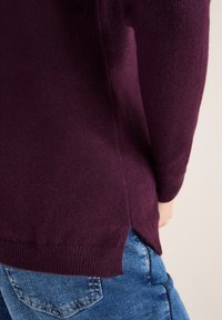 Bordeaux gebreide trui met geribbelde manchetten en een zijsplitontwerp, gecombineerd met blauwe jeans, wat een zachte textuur en casual pasvorm toont.