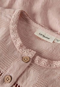 Tessuto morbido rosa con texture, rifinito con pizzo all'uncinetto sul colletto e bottoni di legno, su un capo etichettato "Lil' Atelier."
