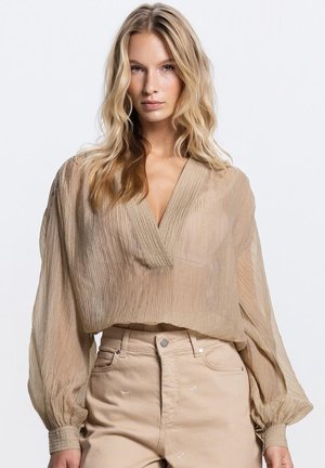 Vrouw met lang blond haar, gekleed in een losse beige geplooide blouse met diepe V-hals en beige broek met hoge taille, tegen een witte achtergrond.