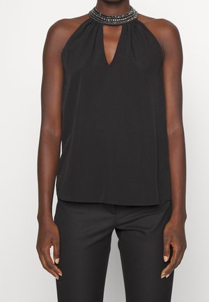 Kobieta w czarnej, bezrękawnikowej bluzce typu halter z koralikowym dekoltem i wycięciem w kształcie kluczyka, w połączeniu z czarnymi spodniami, stojąca na białym tle.