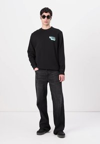 Zwarte sweatshirt met een turkooizen graphic, gecombineerd met donkerdenim jeans. Model draagt zwarte zonnebril en zwarte sneakers. Casual fit.