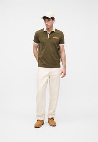 Homme portant un polo vert olive, un pantalon beige, des bottes couleur sable et une casquette de baseball beige, debout devant un fond blanc uni.