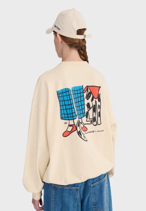Personne portant un sweat-shirt beige avec un graphisme coloré représentant des jambes en pantalon bleu, des chaussures rouges et un chien noir et blanc avec une écharpe rouge dans le dos.