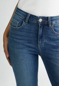 Jeans in denim blu con un design aderente, vita media e tasche frontali. Presentano una cucitura sottile e un bottone in metallo con marchio.