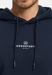 Laivastonsininen huppari pehmeästä kankaasta, jossa on valkoinen etuosan logopainatus, nyörilenkit ja resorit hihansuissa.