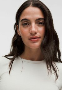 Lichtgekleurde, geribbelde langemouwtop gedragen door een persoon met schouderlang, donker haar en natuurlijke make-up, gecentreerd in het kader.