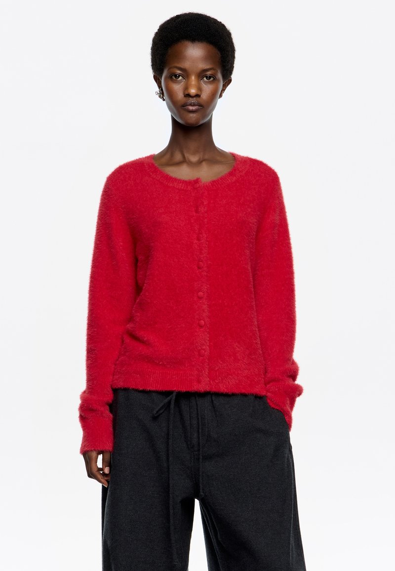 Cardigan rojo de felpa con cuello redondo, cierre frontal de botones y puños de canalé, combinado con pantalones negros sueltos. Textura suave y apariencia cálida.