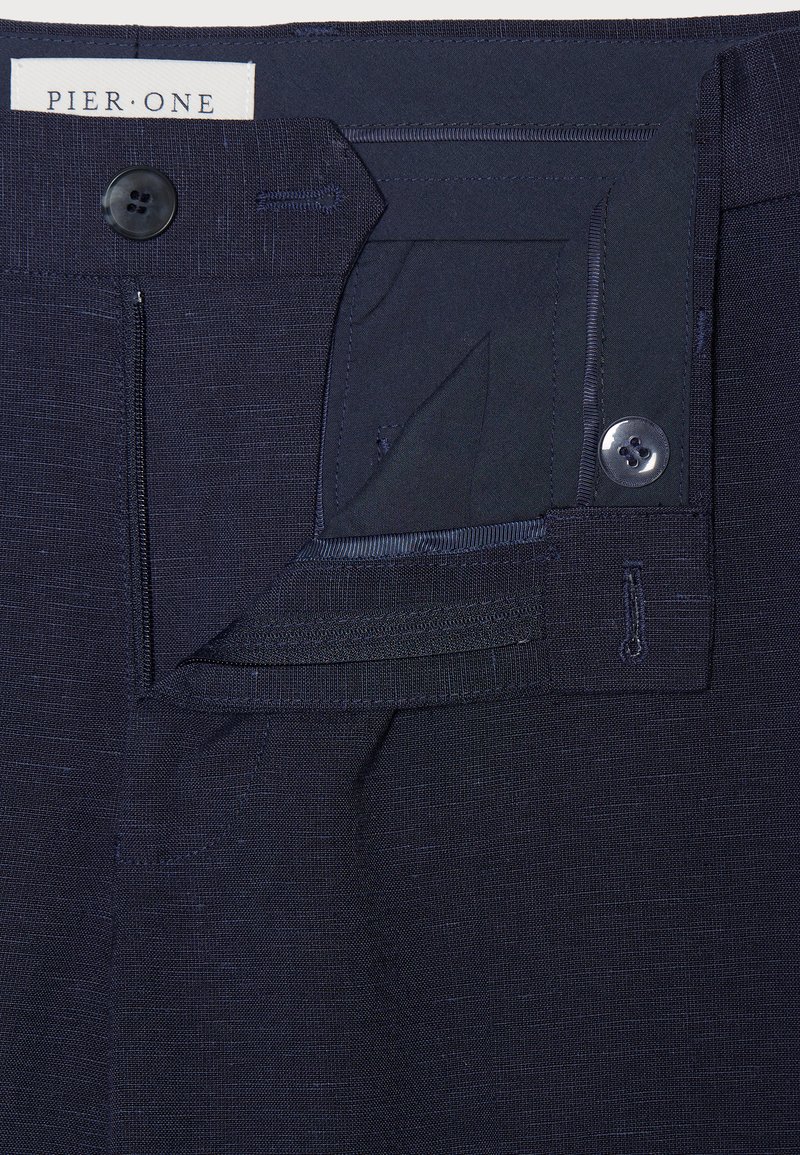 Pantalons bleu marine en tissu texturé. Présentent une fermeture à bouton devant et des détails internes à la taille. Étiquette : "PIER ONE."