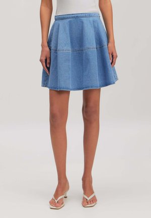 Woman wearing a light blue denim A-line skirt with a white top and white open-toe heeled sandals, standing against a plain background.