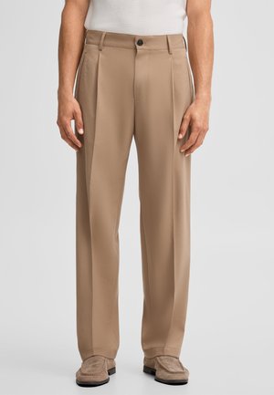 Mann trägt beige plissierte Hose kombiniert mit beigen Wildleder-Loafern und einem weißen, gerippten ärmellosen Oberteil, steht vor einem einfarbigen Hintergrund.