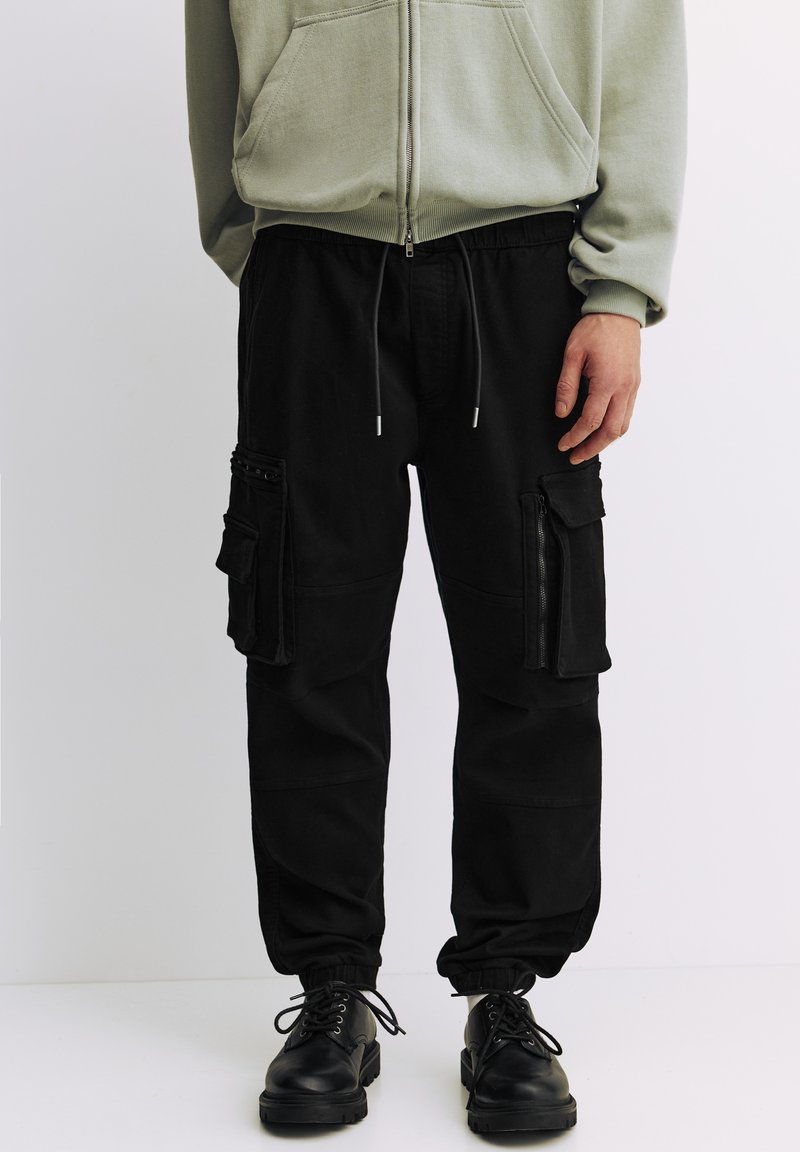 Pantaloni cargo negri cu mai multe buzunare cu fermoar, manșete conice și talie cu șnur, realizați din material durabil.