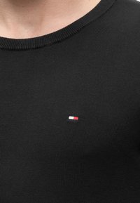 Gros plan d'une personne portant un pull en maille noir avec un petit logo rectangulaire blanc et rouge sur la poitrine.