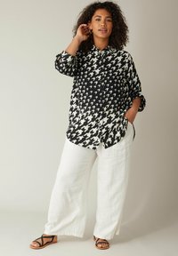 Camisa de patrón de espiga en blanco y negro con cierre de botones y mangas enrolladas, combinada con pantalones de lino blancos y sandalias negras de tiras.