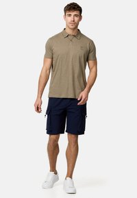 Polo-shirt vert olive avec un logo discret, associé à un short cargo bleu marine avec des poches latérales et une texture douce et tissée. Baskets blanches.