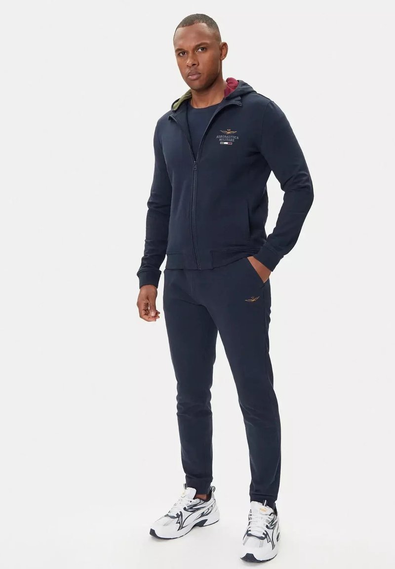 Felpa con zip di colore blu navy con cappuccio interno a contrasto, abbinata a pantaloni jogger blu navy coordinati. Logo in evidenza e dettagli sottili sui capi.
