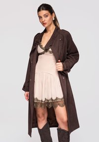 Femme portant une robe légère bordée de dentelle, un manteau long sombre et des bottes hautes sombres, debout devant un fond uni.