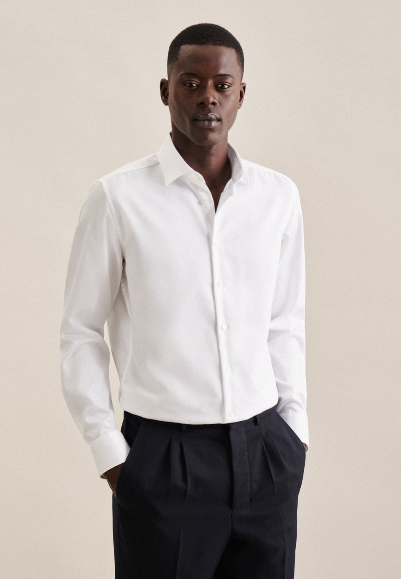 Camicia bianca a maniche lunghe con colletto formale e chiusura a bottoni, abbinata a pantaloni scuri a pieghe, su uno sfondo neutro.