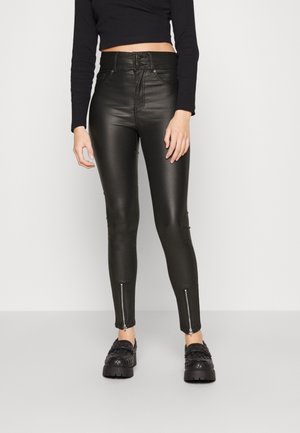 Kobieta nosząca czarne, wysokowycięte spodnie z eko skóry z przodu z guzikami i zamkami przy kostkach, w połączeniu z czarnymi pikowanymi butami na platformie.