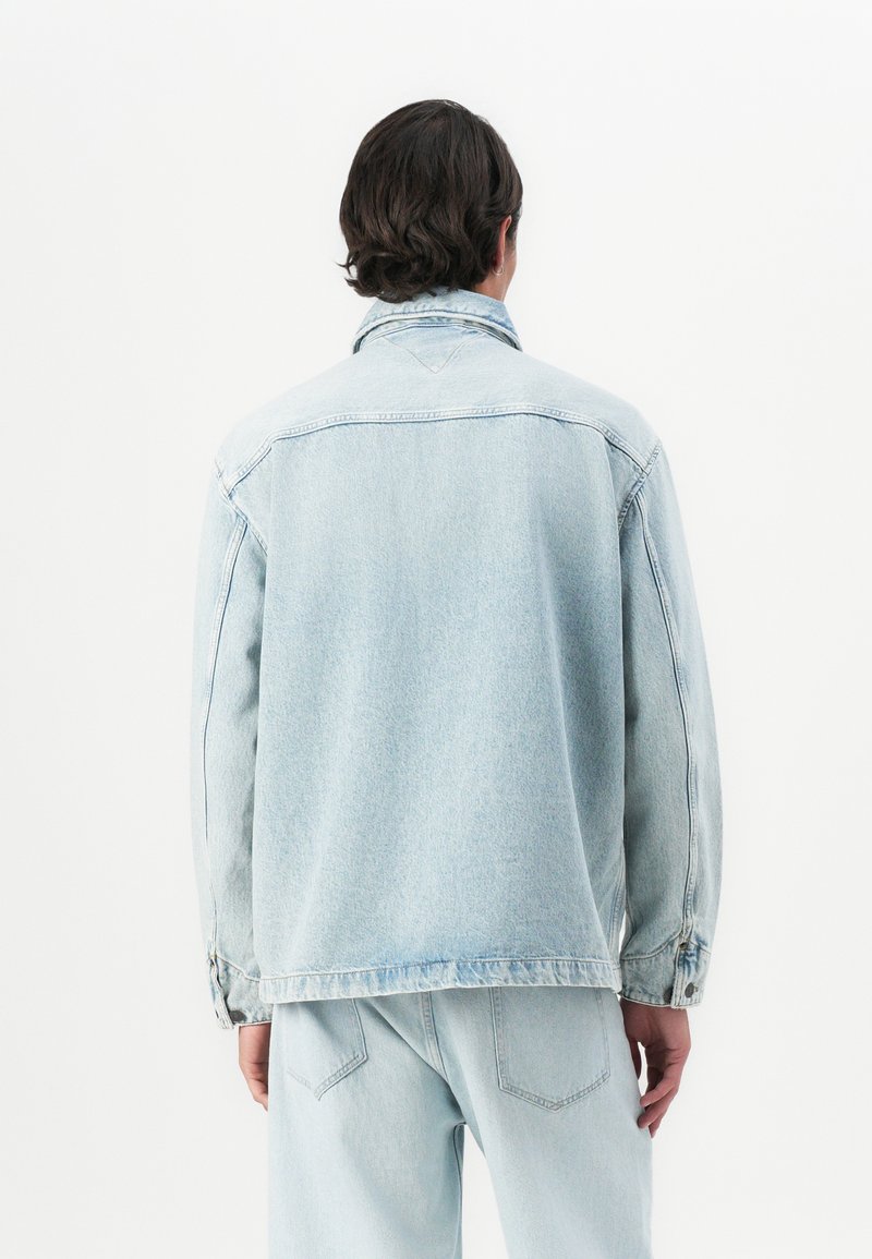 Personne avec des cheveux courts et foncés portant une veste en denim bleu clair et un jean assorti, se tenant de dos contre un fond blanc.