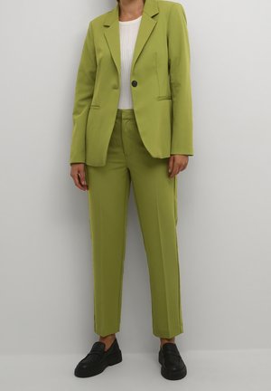 Personne portant un costume sur mesure vert clair avec un bouton noir, associé à un top blanc et des mocassins noirs, debout contre un mur uni.