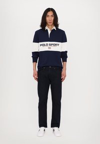 Navy un balts garām piedurknēm polo krekls ar "POLO SPORT" logotipu, apvienots ar tumšo džinsu biksēm un baltām sporta apaviem. Modelis stāv pretī vienkāršai fona.