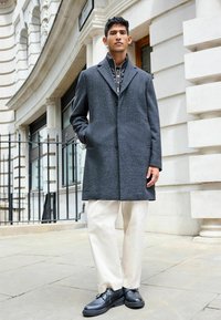 Palton gri în model herringbone cu rever în formă de v, buzunare laterale, purtat peste o cămașă cu un model și asortat cu pantaloni crema și pantofi negri.