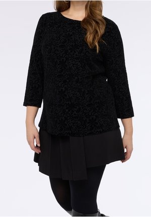 Femme portant un haut noir en velours à manches longues à motif floral, associé à une jupe plissée noire, des leggings noirs et des bottes montantes.