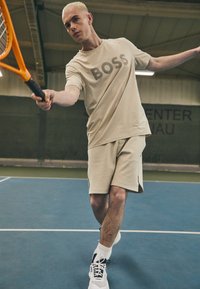 Camiseta deportiva beige con el logo "BOSS", combinada con pantalones cortos a juego. El jugador sostiene una raqueta de tenis naranja sobre una superficie de pista azul.