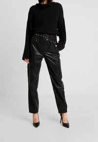 Un pull noir à côtes avec un col haut, associé à un pantalon cargo noir brillant avec des poches et des poignets resserrés. Des escarpins noirs à talons aiguilles.