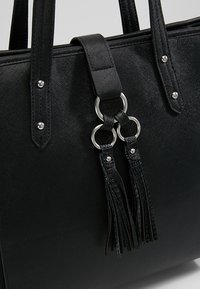 Czarna torebka na ramię o teksturowanej powierzchni, z dwoma uchwytami, srebrnymi detalami z metalu oraz z ozdobnym frędzlem i elementami z faux skóry.