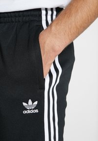 Svarta träningbyxor med vitt tre-randsdesign på sidorna. Har en elastisk midja och en broderad vit Adidas-logotyp på framsidan.