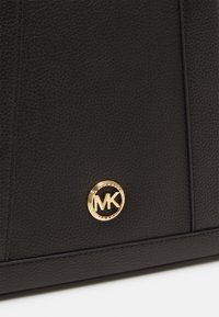 Sac en cuir noir à texture granuleuse avec un badge logo circulaire doré à l'avant, présentant des détails cousus et une texture lisse.