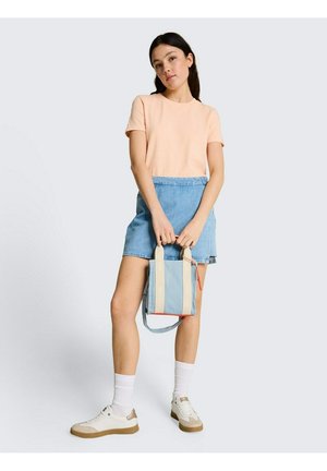Jonge vrouw in perzikkleurig T-shirt en blauwe denim rok die een lichtblauwe draagtas vasthoudt, draagt witte sokken en beige sneakers, staand tegen een effen achtergrond.