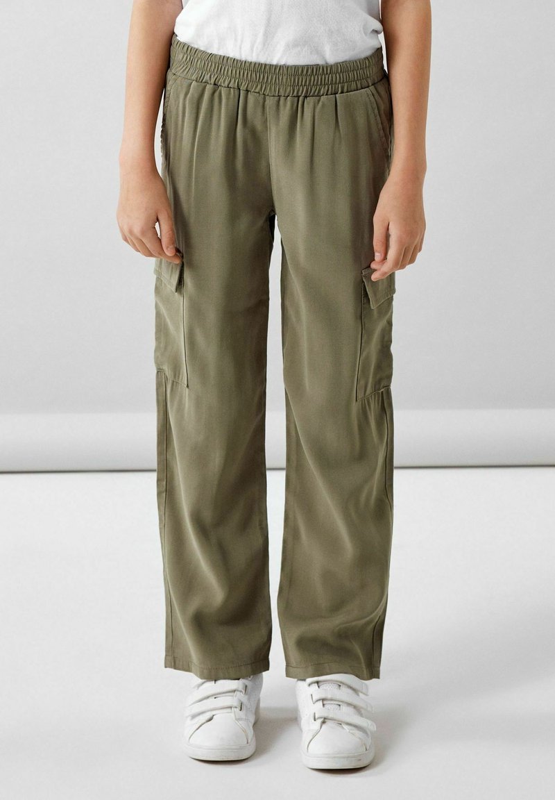 Enfant portant un pantalon cargo vert olive avec une ceinture élastique et des baskets blanches à scratch, debout devant un fond uni.