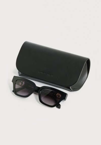 Μαύρα ορθογώνια γυαλιά ηλίου με σκούρους φακούς τοποθετημένα μπροστά από μια αντίστοιχη μαύρη θήκη Giorgio Armani πάνω σε λευκή επιφάνεια.