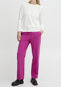 Femme portant un pull blanc à manches longues, un pantalon rose vif à jambe droite et des mocassins noirs épais, se tenant avec une main dans sa poche.