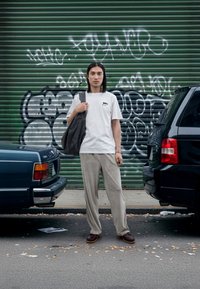 White t-shirt with a small graphic, grey wide-leg trousers, brown shoes, and a grey tote bag. Background features a green metal shutter and graffiti.