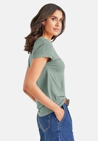 Groen T-shirt met korte mouwen en een ronde hals, gemaakt van zacht materiaal. Gecombineerd met blauwe high-waisted jeans, waarbij het zijprofiel en het gestructureerde haar zichtbaar zijn.