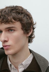 Nærbillede af en person med krøllet brunt hår, iført en stribet skjorte under en mørk krave, med glat hud og neutral baggrund.